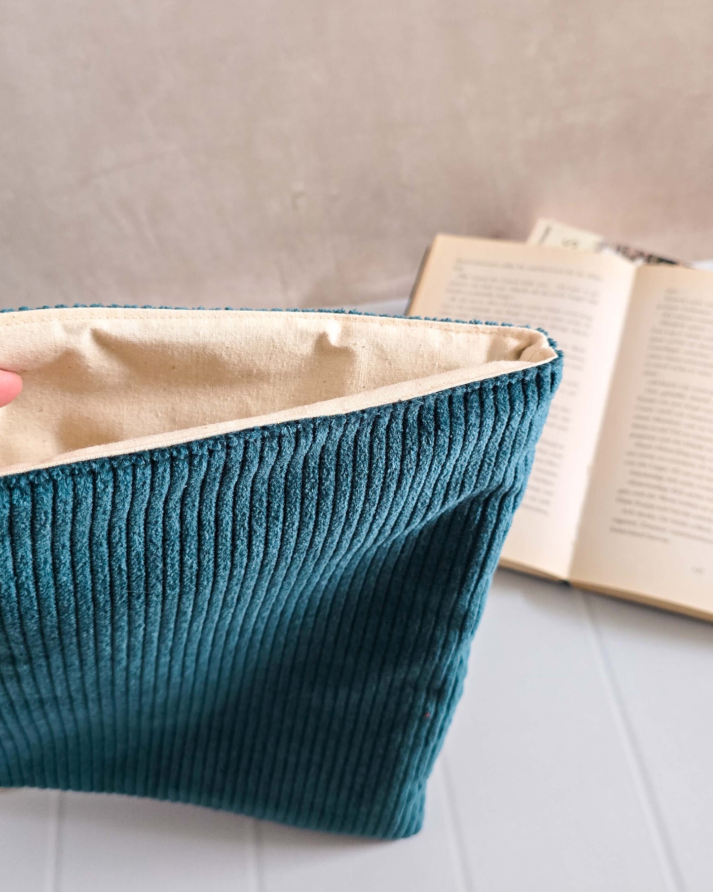 Handgenähte Buchhülle in dunkelpetrolfarbenem Cord vor aufgeschlagenem Buch und Pflanze