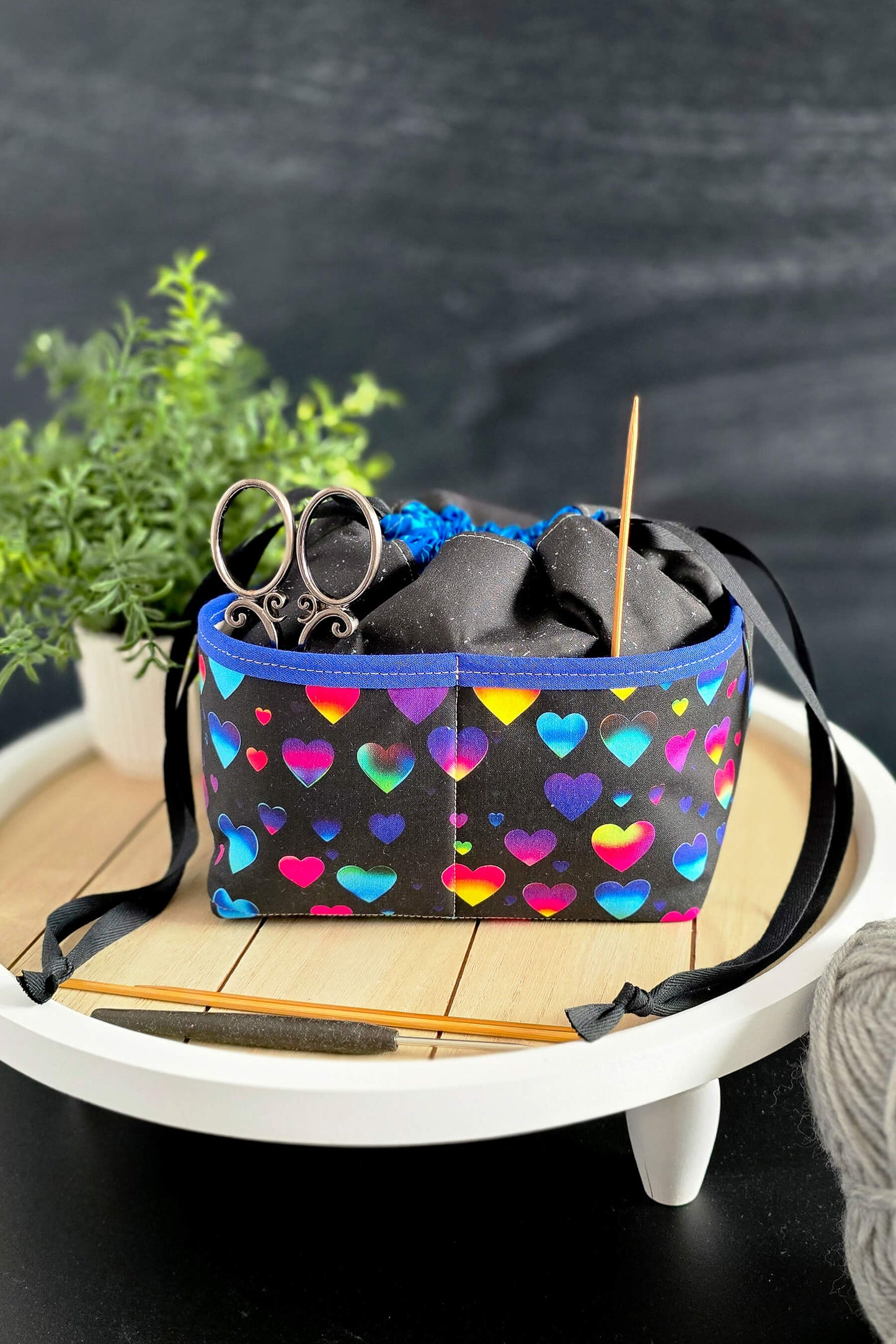 bunte Projekttasche mit Herzen und Kordelzug, gefüllt mit Strickzubehör, steht auf einem Holztisch