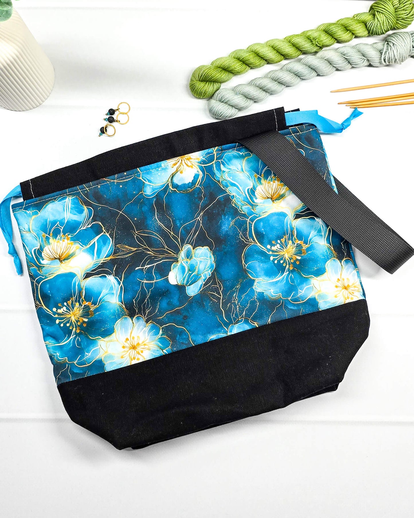 Projekttasche mit Kordelzug und schwarzem Tragegurt aus türkis-blauem Blumenstoff mit Goldakzenten und schwarzem Boden. Bietet Platz für 400g Wolle und schützt das Strickzeug vor Verschmutzung. Handgenäht in Bayern von Nadelmikado.