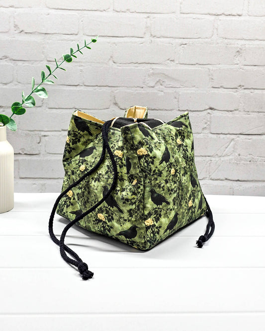 Mittlere grüne Projekttasche mit Kordelzug im Rice Bag Stil, gemustert mit Raben und Rosen, genutzt als Strickbeutel für ein Tuchprojekt mit bis zu 400g Wolle. Handgenäht in Bayern von Nadelmikado.