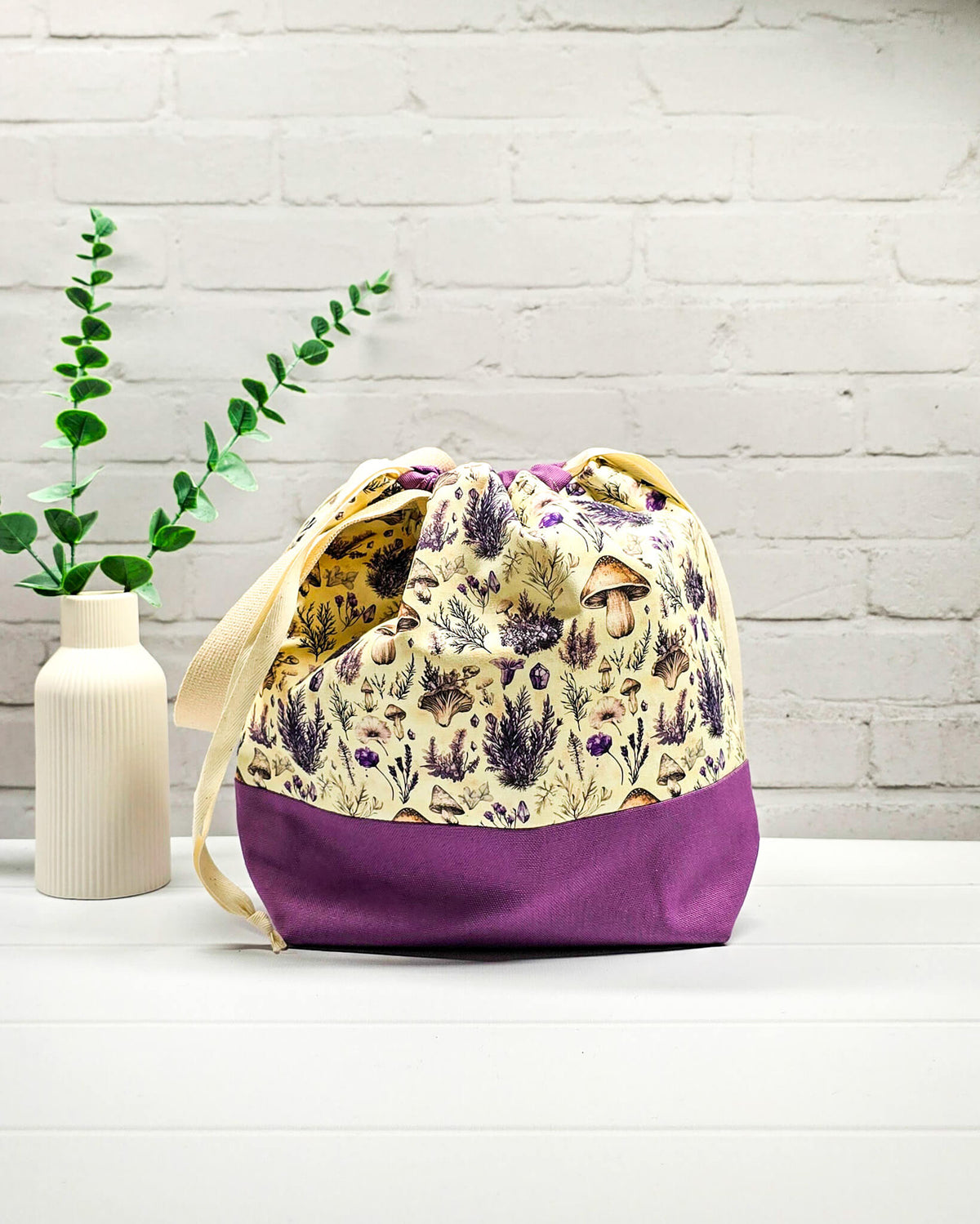 Strickbeutel Größe M mit Pilzen, Kräutern und violetten Blüten auf hellem Grund sowie lilafarbenem Boden und Kordelzug. Handgenäht in Bayern von Nadelmikado.