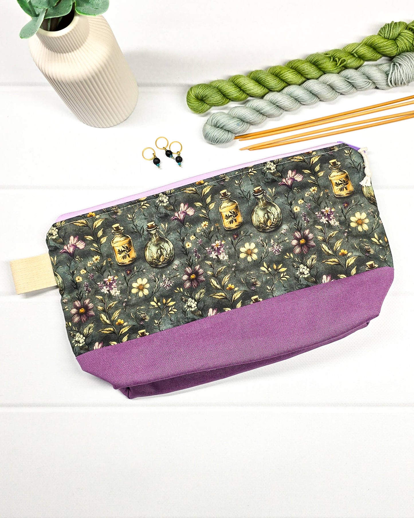 Projekttasche Isvind S mit Elixierfläschchen und Wildblumen auf dunkelgrünem Grund, lilafarbenem Canvas-Boden und lila Reißverschluss für Sockenwolle. Handgenäht in Bayern von Nadelmikado.