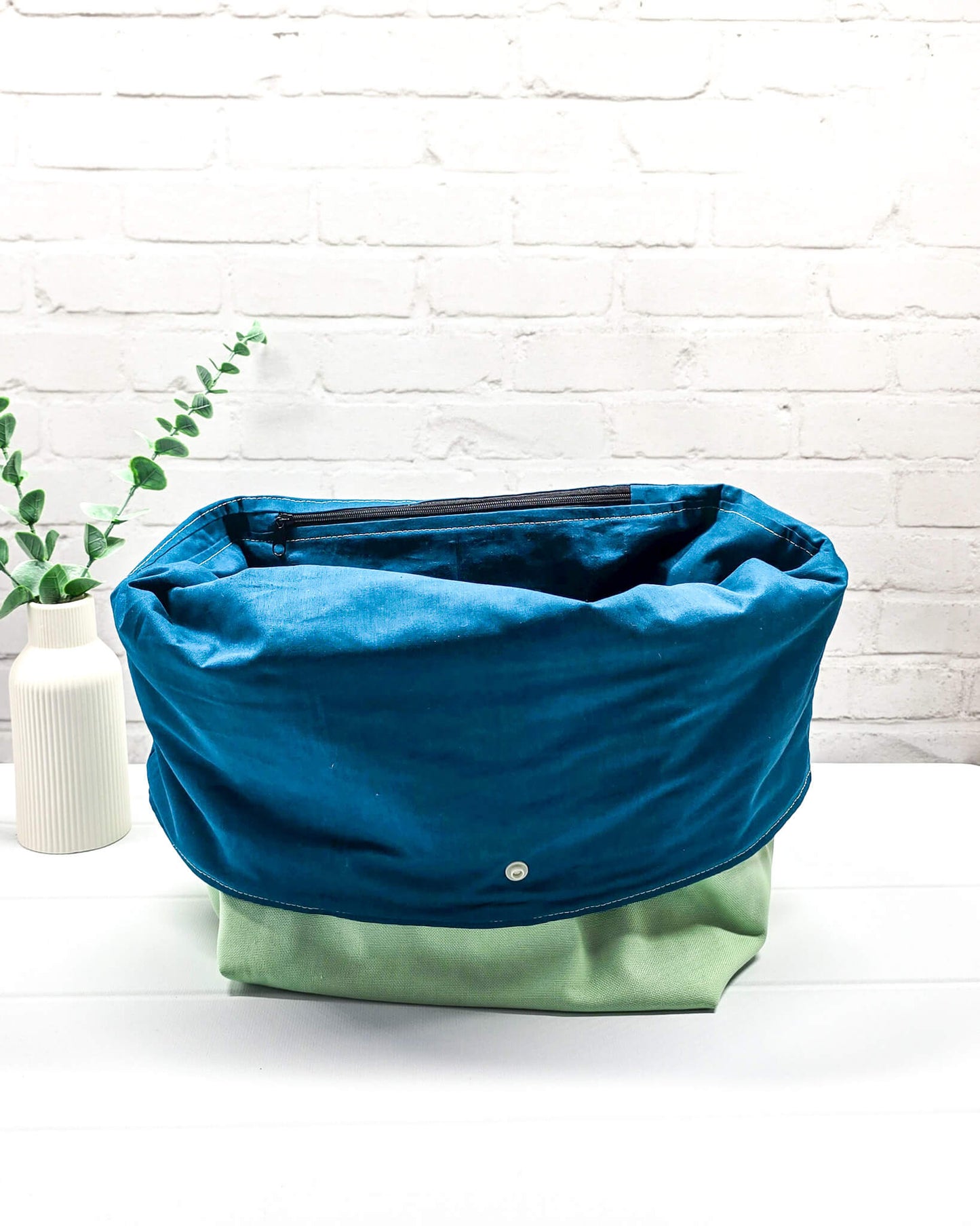 Projekttasche Rika im Roll-Top-Design aus hellgrünem Canvas mit vertikalem, dunkelgrünem Schriftzug „KNITTING“ und petrolfarbenem Gurtband-Verschluss. Handgenäht in Bayern von Nadelmikado.