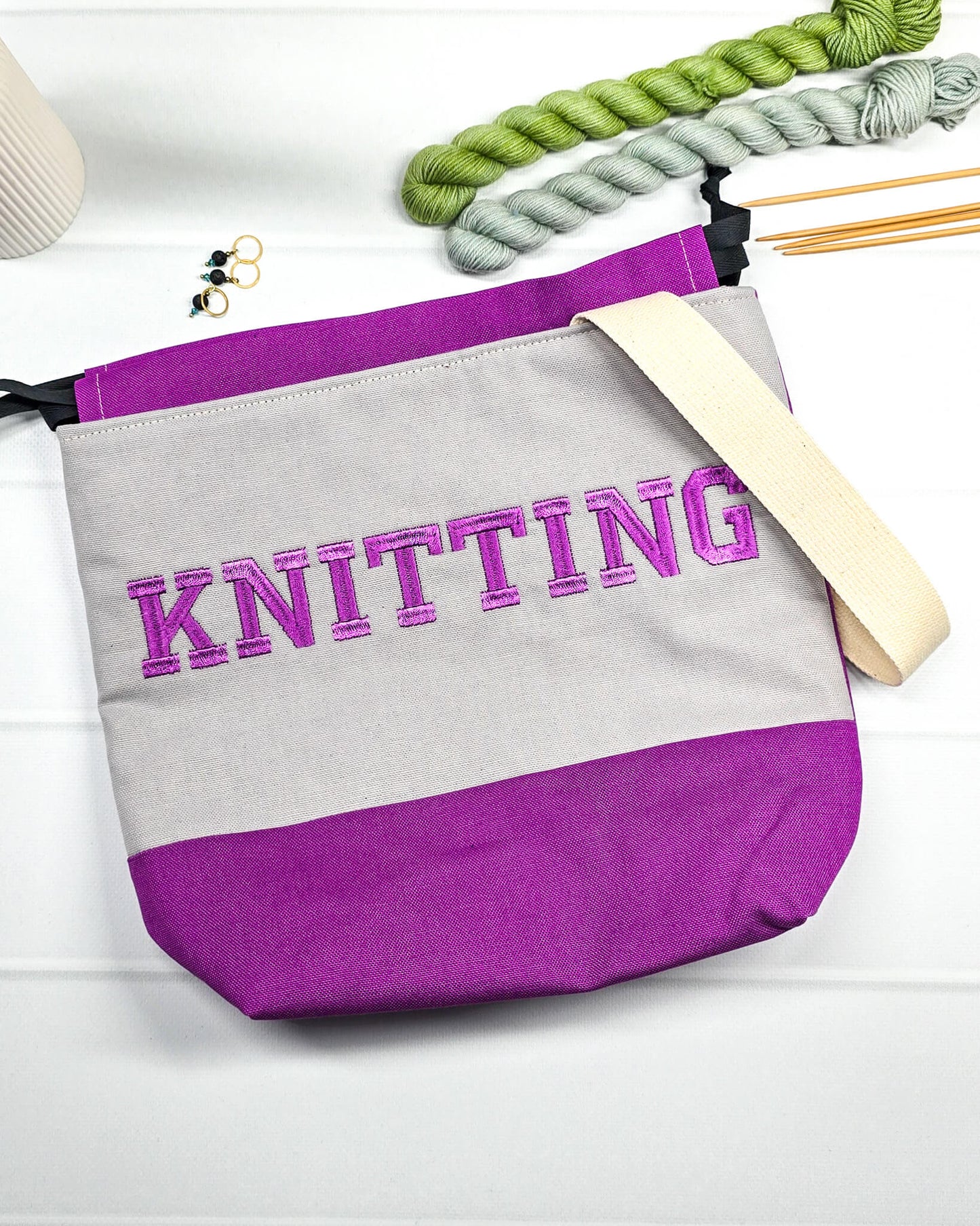 Bestickte Projekttasche Tuva M aus grauem und lila Canvas-Stoff mit violettem Schriftzug KNITTING, schwarzem Kordelzug und cremefarbenem Tragegurt. Bietet Platz für 400g Wolle und schützt das Strickzeug vor Verheddern. Handgenäht in Bayern von Nadelmikado.