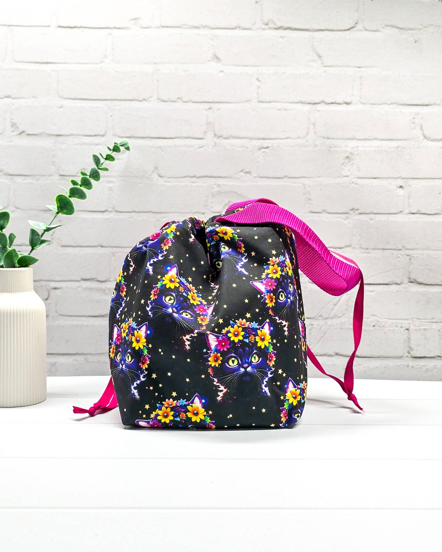 Projekttasche Tuva M aus schwarzem Baumwollstoff mit bunten Blumen-Katzen, pinkem Kordelzug und pinker Trageschlaufe. Handgenäht in Bayern von Nadelmikado.