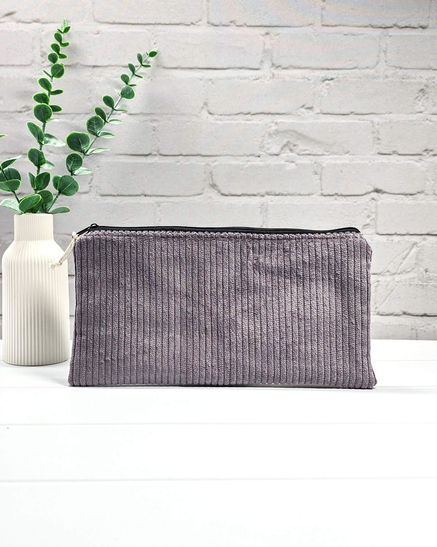 Kleine graulila Reißverschlusstasche aus Cord-Stoff für Strickzubehör, liegend auf weißem Untergrund mit einer Schere, Garnsträngen und Holznadeln. Handgenäht in Bayern von Nadelmikado.