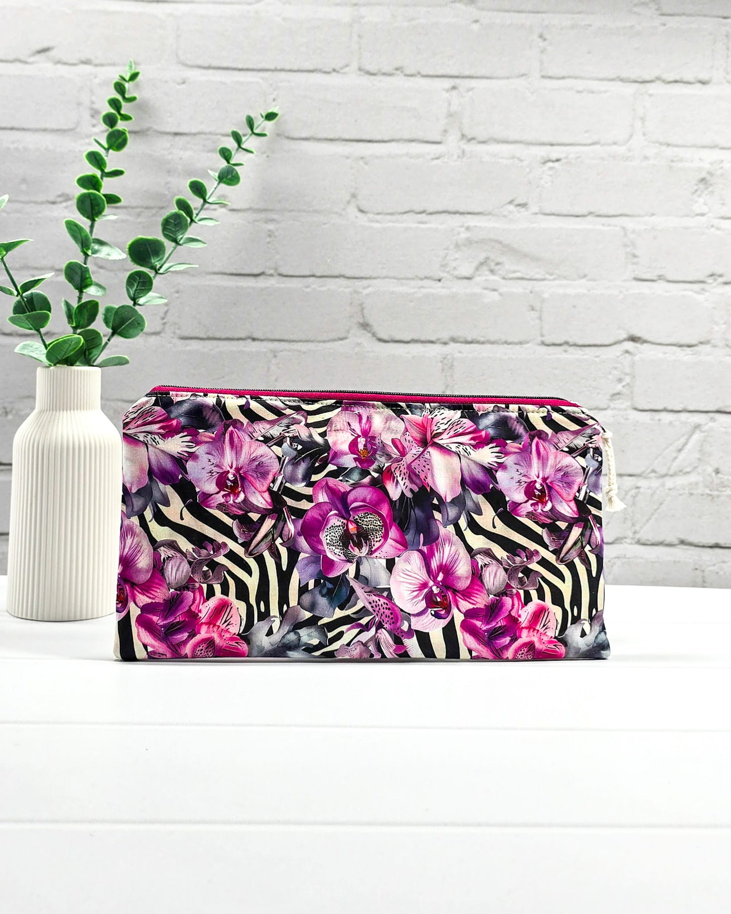 Längliche Tasche mit pinkfarbenem Reißverschluss, hellem Kordel-Zipper und auffälligem Muster aus lila Orchideenblüten über einem schwarz-weißen Zebramuster. Handgenäht in Bayern von Nadelmikado.