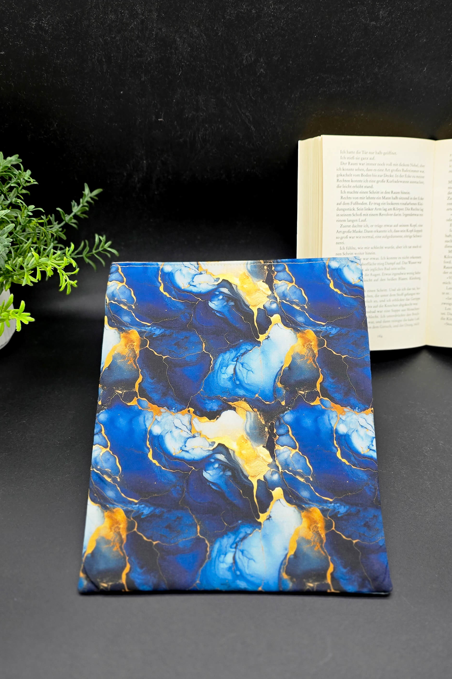 Handgenähte Buchhülle mit blau-goldenem Marmormuster, liegend auf schwarzem Hintergrund, Vorderansicht