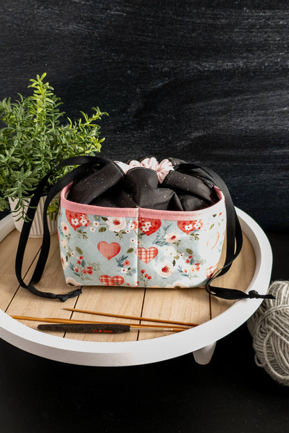 Handarbeitstasche liebevoll platziert auf einem Tisch, mit Garnknäuel und Stricknadeln daneben.