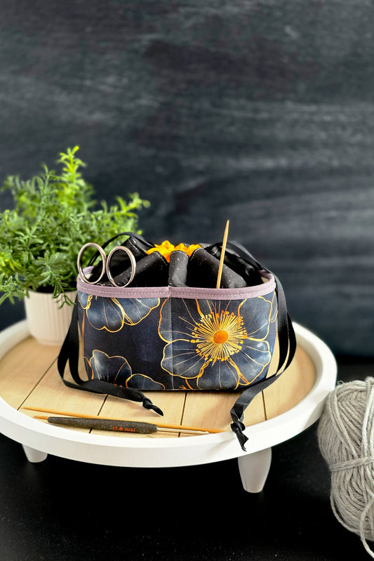 handgenähte Projekttasche mit Blumenmuster, gefüllt mit Strickzubehör, steht auf einem Holztisch vor dunklem Hintergrund