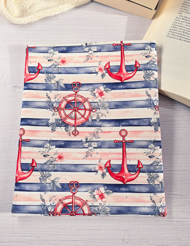 Maritime Buchhülle mit Anker-Muster in Blau-Rot-Weiß auf einer flachen Oberfläche präsentiert.