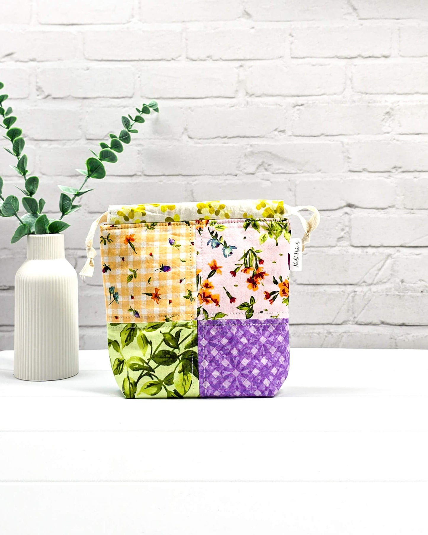 Kleine Projekttasche XS aus weißem Baumwollstoff mit einem bunten Blumen-Muster, einem schwarzen, weiß gepunkteten oberen Rand und schwarzen Kordelzügen. Handgenäht in Bayern von Nadelmikado. 