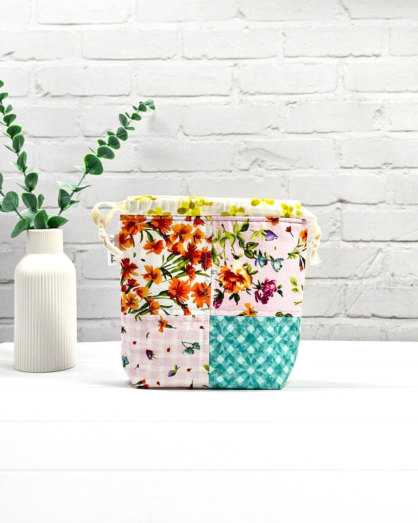 Kleine Projekttasche XS aus weißem Baumwollstoff mit einem bunten Blumen-Muster, einem schwarzen, weiß gepunkteten oberen Rand und schwarzen Kordelzügen. Handgenäht in Bayern von Nadelmikado. 