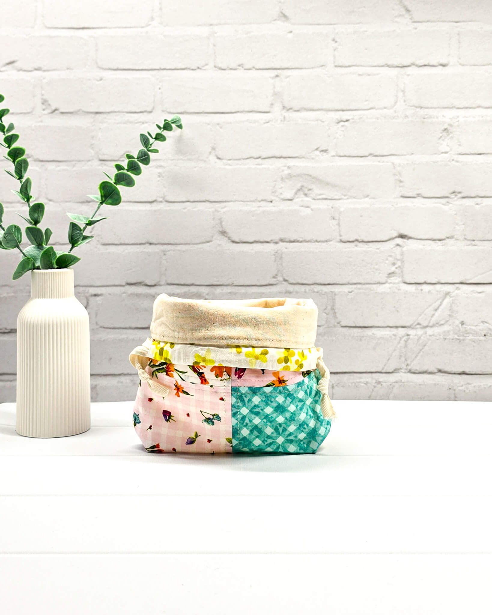 Kleine Projekttasche XS aus weißem Baumwollstoff mit einem bunten Blumen-Muster, einem schwarzen, weiß gepunkteten oberen Rand und schwarzen Kordelzügen. Handgenäht in Bayern von Nadelmikado. 