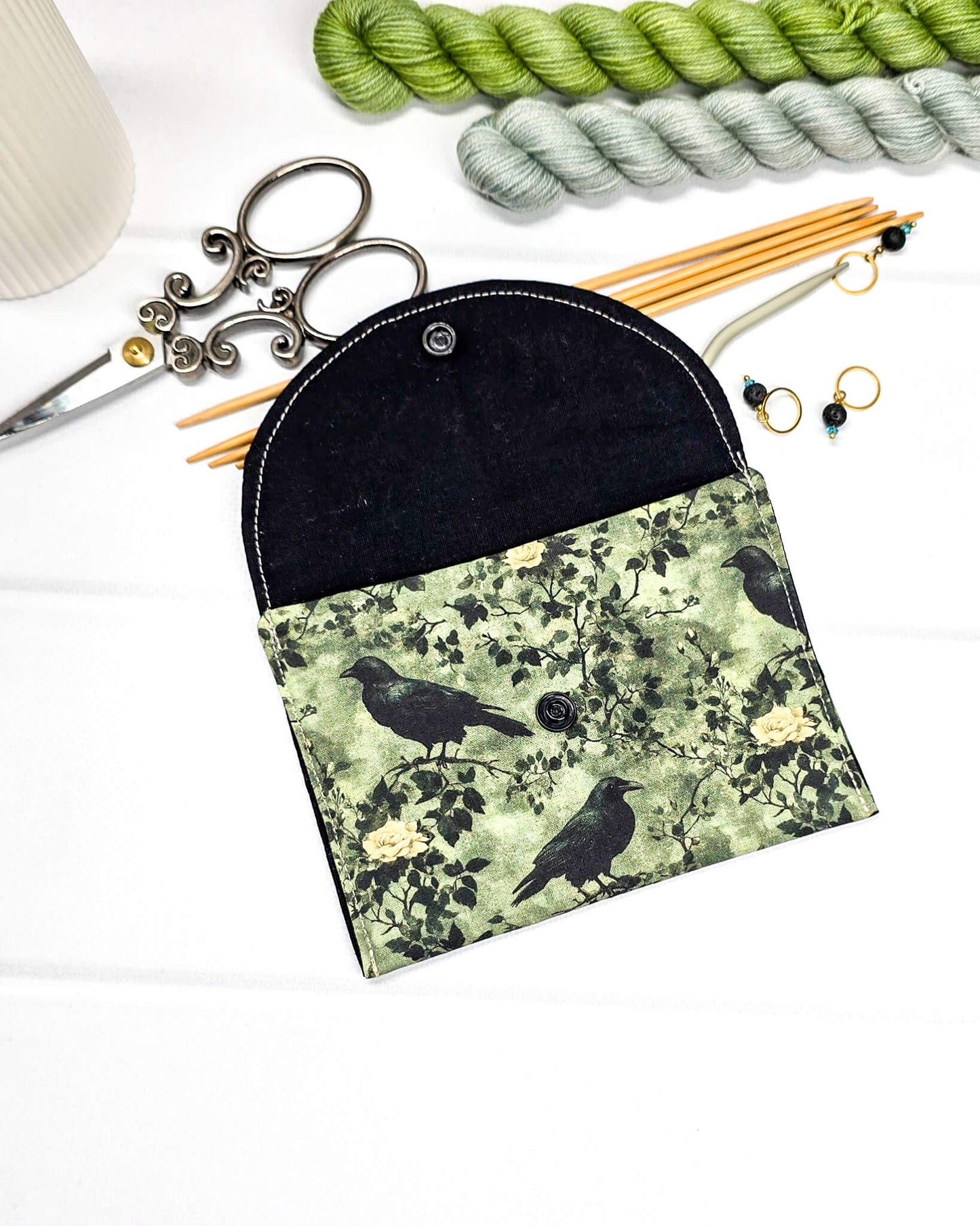 Kleine grüne Zubehörtasche mit Druckknopf, gemustert mit Raben und Rosen, genutzt als Mini Pouch für Maschenmarkierer und Strickzubehör. Handgenäht von Nadelmikado.