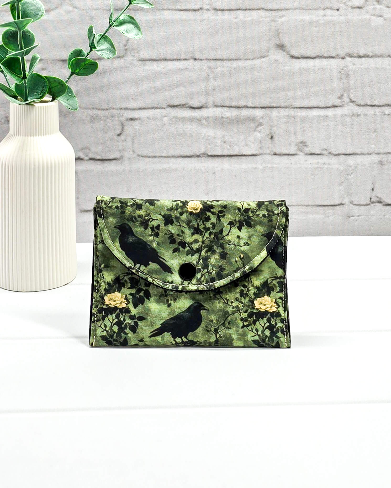 Kleine grüne Zubehörtasche mit Druckknopf, gemustert mit Raben und Rosen, genutzt als Mini Pouch für Maschenmarkierer und Strickzubehör. Handgenäht von Nadelmikado.