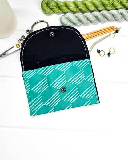 Kleine, flache Mini-Pouch für Strickzubehör mit hellem, grafischem Linienmuster auf smaragdgrünem Stoff, verschlossen mit einer Klappe und einem schwarzen Druckknopf. Handgenäht in Bayern von Nadelmikado.