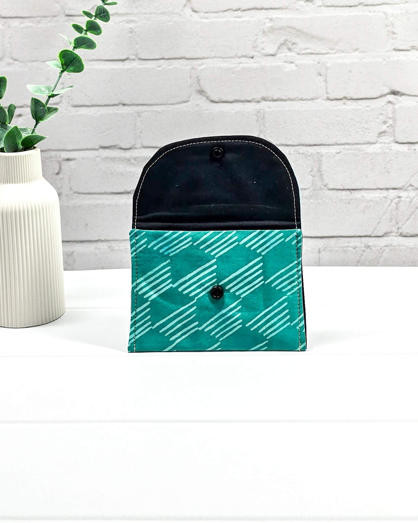 Kleine, flache Mini-Pouch für Strickzubehör mit hellem, grafischem Linienmuster auf smaragdgrünem Stoff, verschlossen mit einer Klappe und einem schwarzen Druckknopf. Handgenäht in Bayern von Nadelmikado.