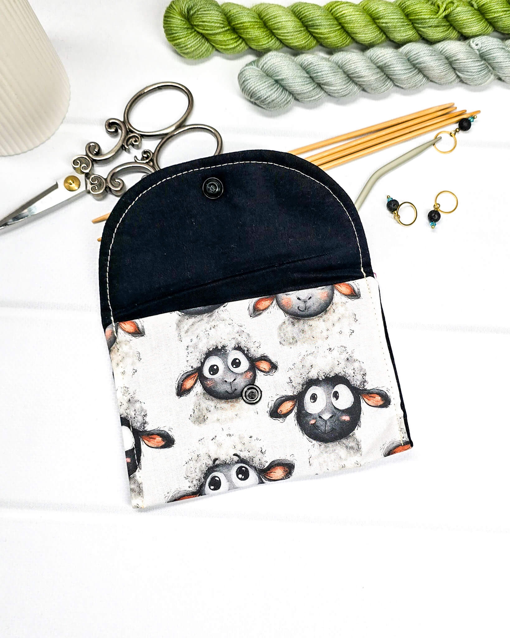 Kleine, flache Mini-Pouch für Handarbeitszubehör mit drolligem Schaf-Motiv auf hellgrau meliertem Stoff, verschlossen mit einer Klappe und einem schwarzen Druckknopf. Handgenäht in Bayern von Nadelmikado. 