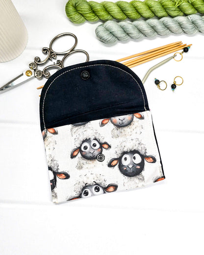 Kleine, flache Mini-Pouch für Handarbeitszubehör mit drolligem Schaf-Motiv auf hellgrau meliertem Stoff, verschlossen mit einer Klappe und einem schwarzen Druckknopf. Handgenäht in Bayern von Nadelmikado. 
