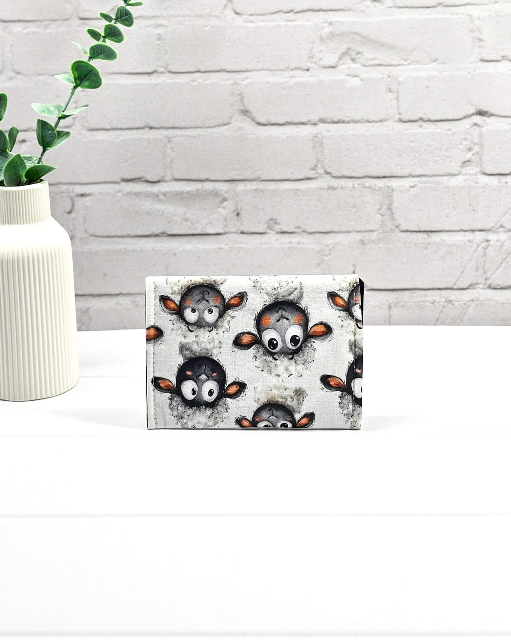 Kleine, flache Mini-Pouch für Handarbeitszubehör mit drolligem Schaf-Motiv auf hellgrau meliertem Stoff, verschlossen mit einer Klappe und einem schwarzen Druckknopf. Handgenäht in Bayern von Nadelmikado. 