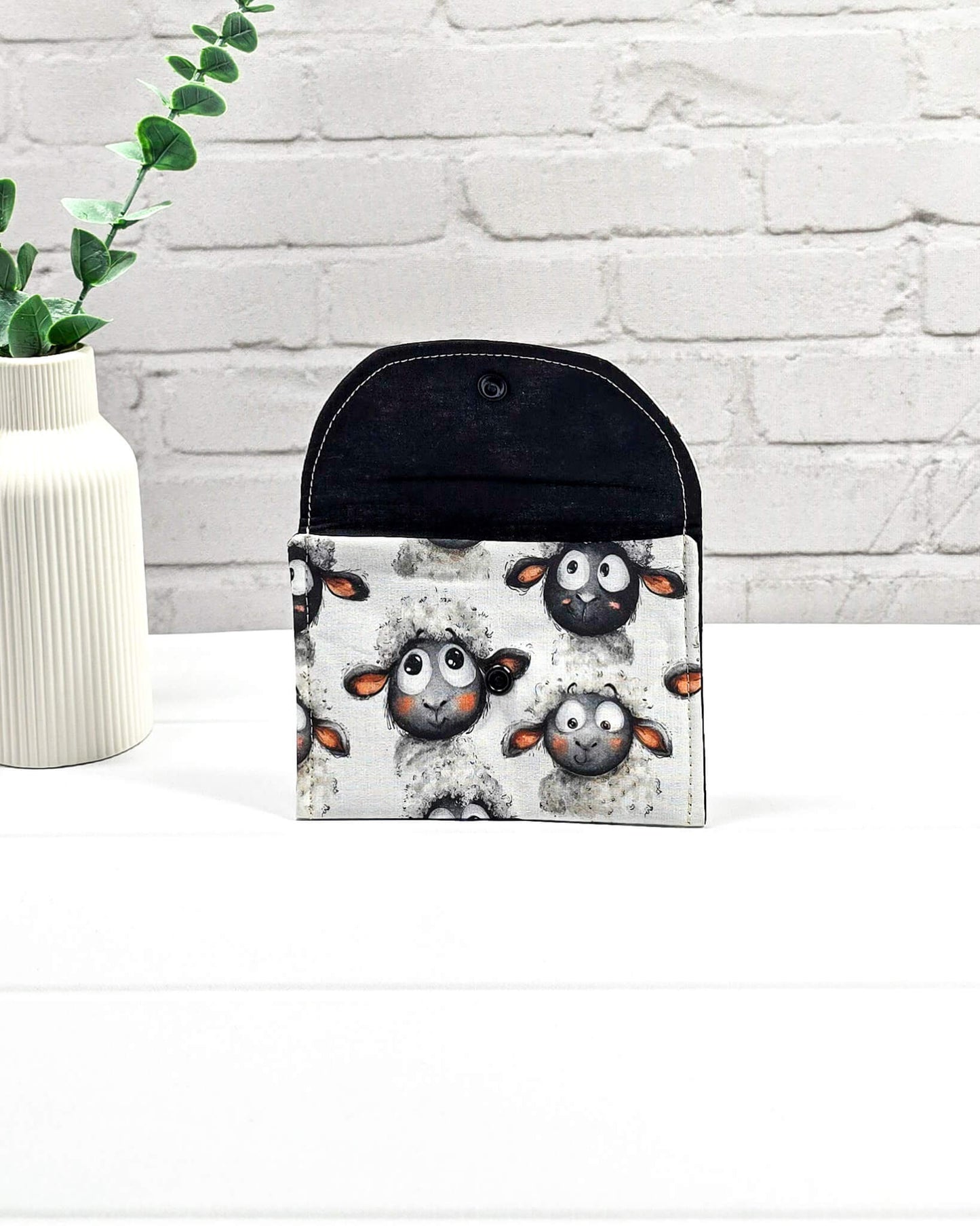 Kleine, flache Mini-Pouch für Handarbeitszubehör mit drolligem Schaf-Motiv auf hellgrau meliertem Stoff, verschlossen mit einer Klappe und einem schwarzen Druckknopf. Handgenäht in Bayern von Nadelmikado. 