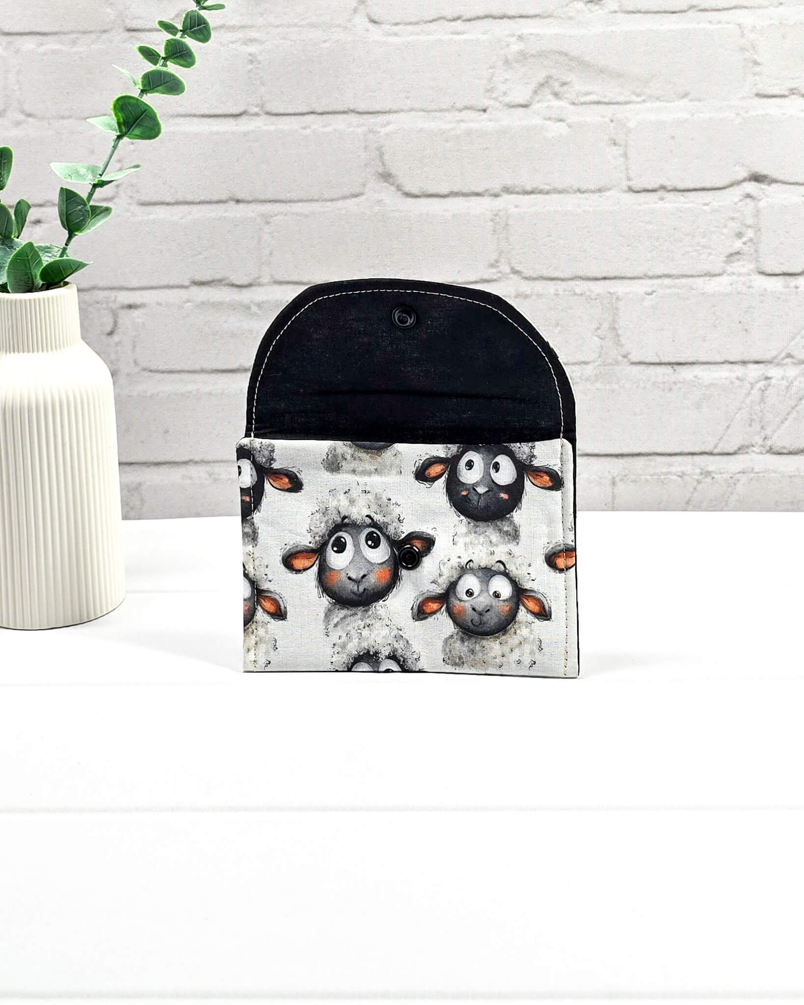 Kleine, flache Mini-Pouch für Handarbeitszubehör mit drolligem Schaf-Motiv auf hellgrau meliertem Stoff, verschlossen mit einer Klappe und einem schwarzen Druckknopf. Handgenäht in Bayern von Nadelmikado. 
