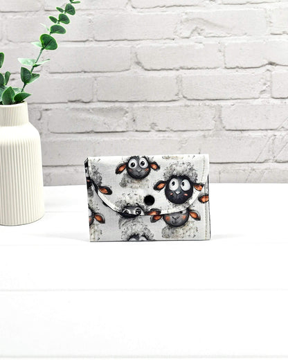 Kleine, flache Mini-Pouch für Handarbeitszubehör mit drolligem Schaf-Motiv auf hellgrau meliertem Stoff, verschlossen mit einer Klappe und einem schwarzen Druckknopf. Handgenäht in Bayern von Nadelmikado. 