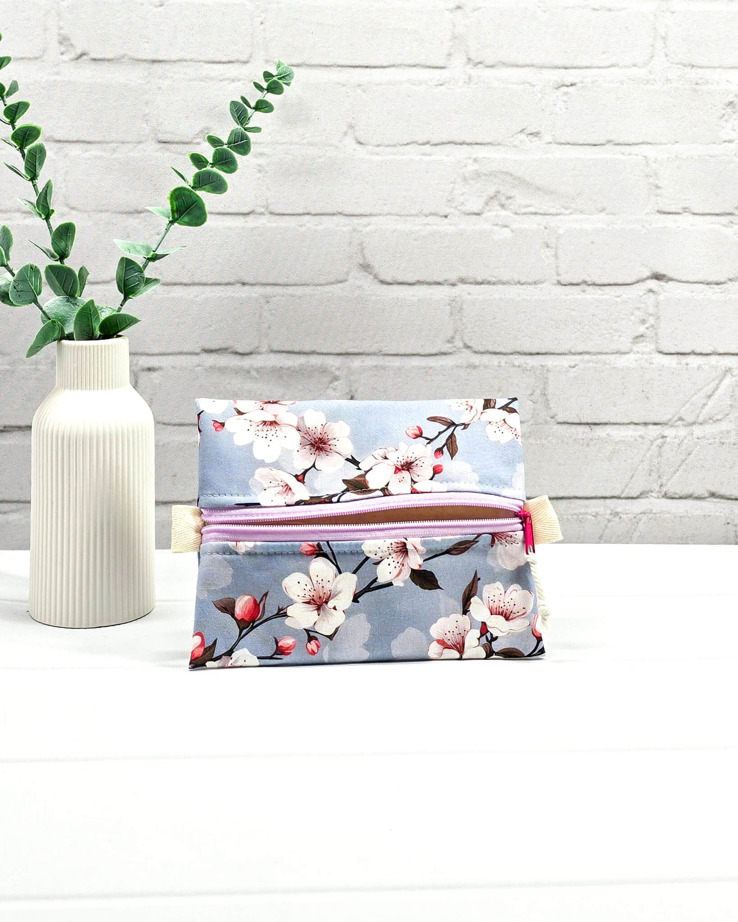 Mini Zip Pouch mit einem Kirschblüten-Muster auf taubenblauem Stoff, einem hellvioletten mittigen Reißverschluss und hellen Stofflaschen. Handgenäht in Bayern von Nadelmikado. 