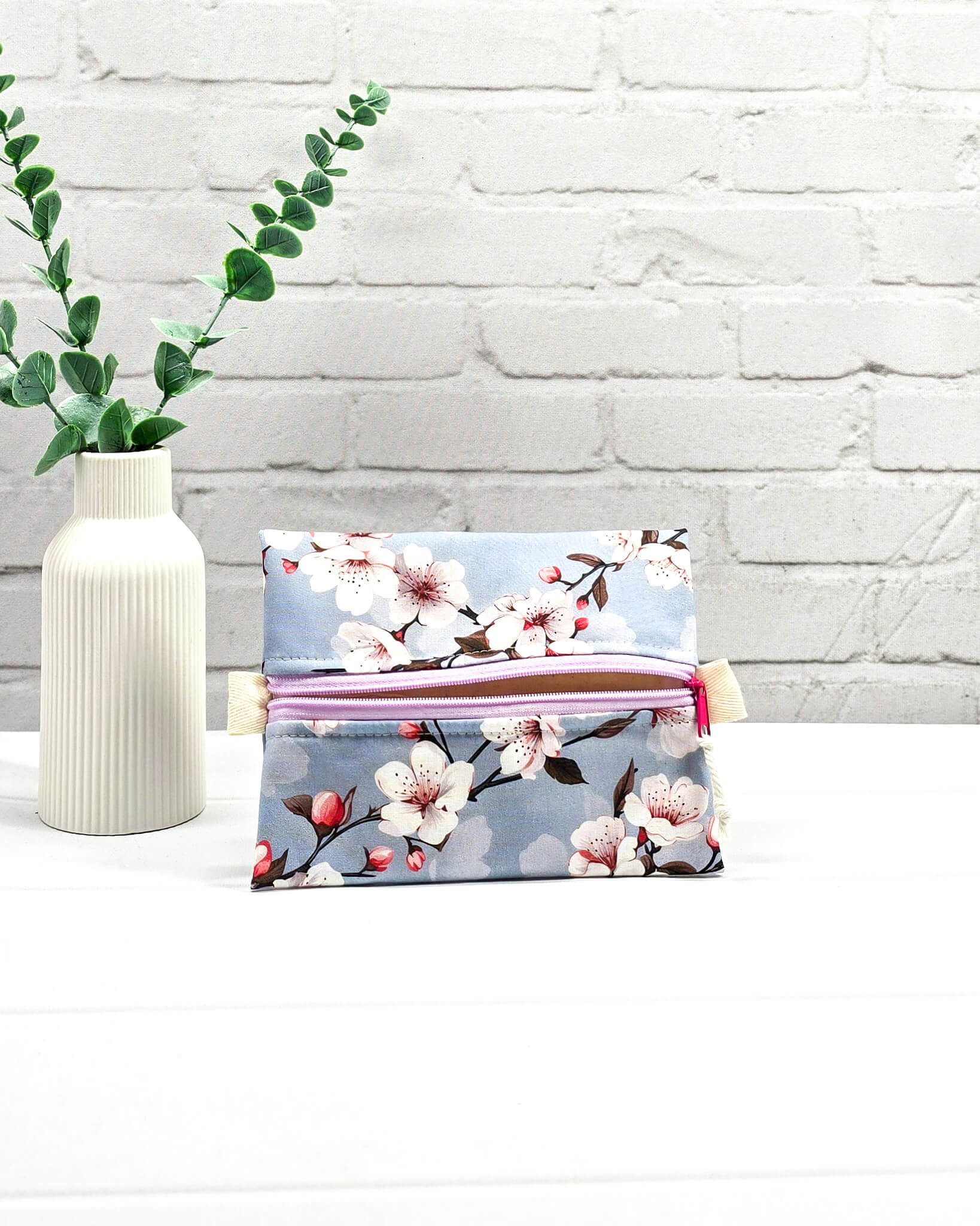 Mini Zip Pouch mit einem Kirschblüten-Muster auf taubenblauem Stoff, einem hellvioletten mittigen Reißverschluss und hellen Stofflaschen. Handgenäht in Bayern von Nadelmikado. 