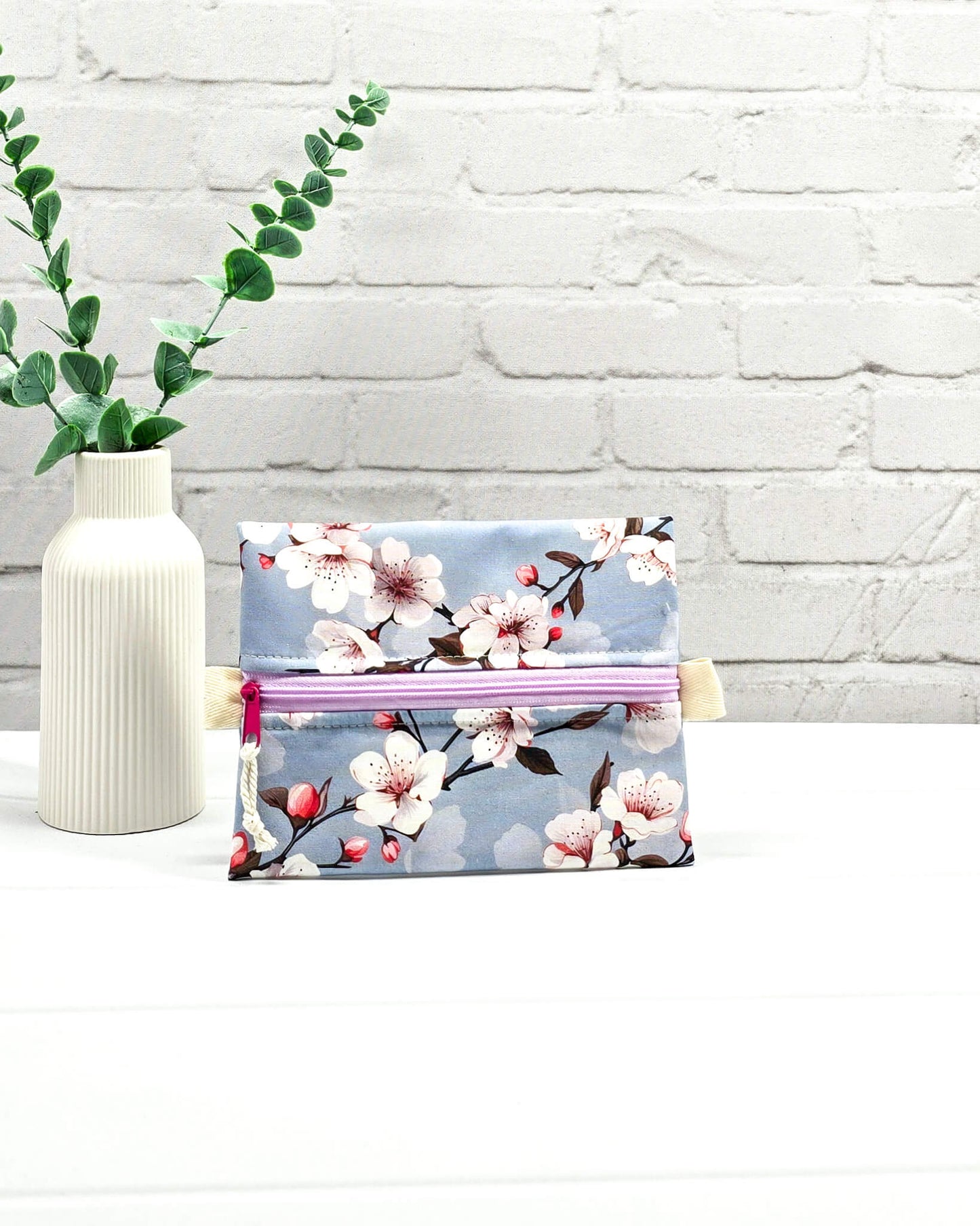 Mini Zip Pouch mit einem Kirschblüten-Muster auf taubenblauem Stoff, einem hellvioletten mittigen Reißverschluss und hellen Stofflaschen. Handgenäht in Bayern von Nadelmikado. 