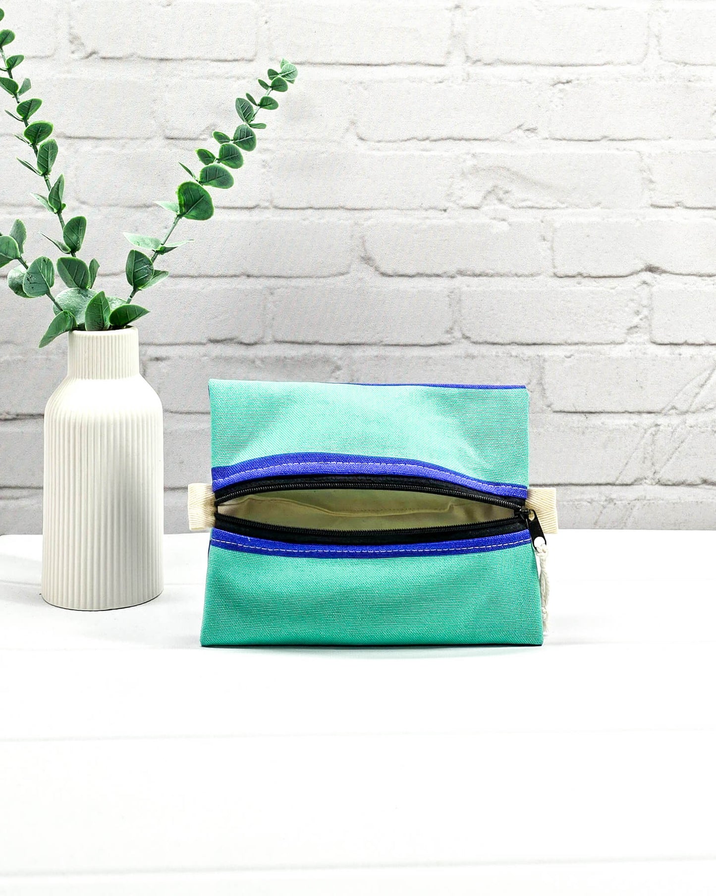 Mini Zip Pouch in Mintgrün mit einem blauen Kontraststreifen, einem schwarzen mittigen Reißverschluss und hellen Stofflaschen. Handgenäht in Bayern von Nadelmikado. 