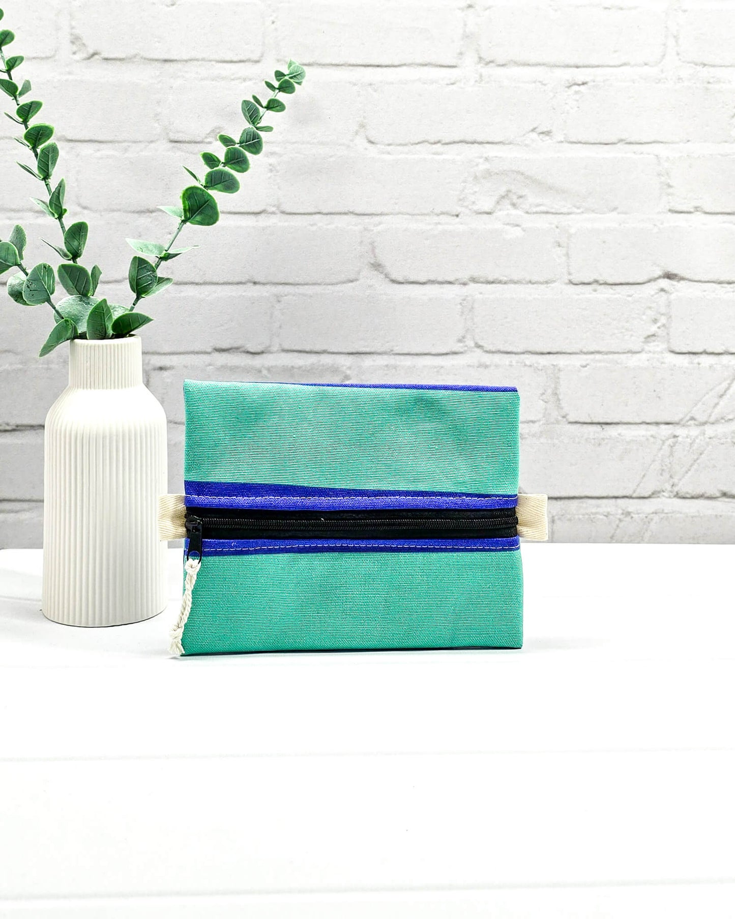 Mini Zip Pouch in Mintgrün mit einem blauen Kontraststreifen, einem schwarzen mittigen Reißverschluss und hellen Stofflaschen. Handgenäht in Bayern von Nadelmikado. 
