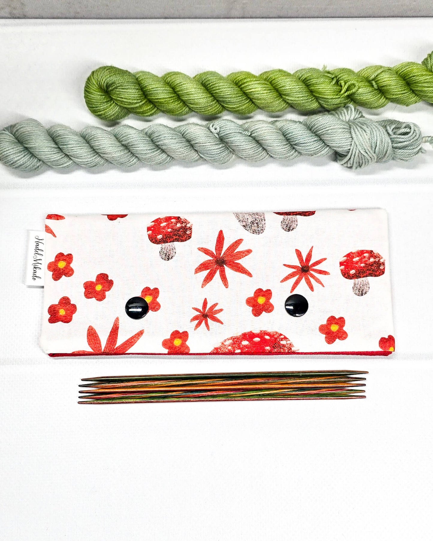 Nadelgarage für 15 cm lange Nadelspiele mit zwei schwarzen Druckknöpfen, roten Fliegenpilzen und herbstlichen Blumenmotiven auf hellem Stoff. Handgenäht in Bayern von Nadelmikado.