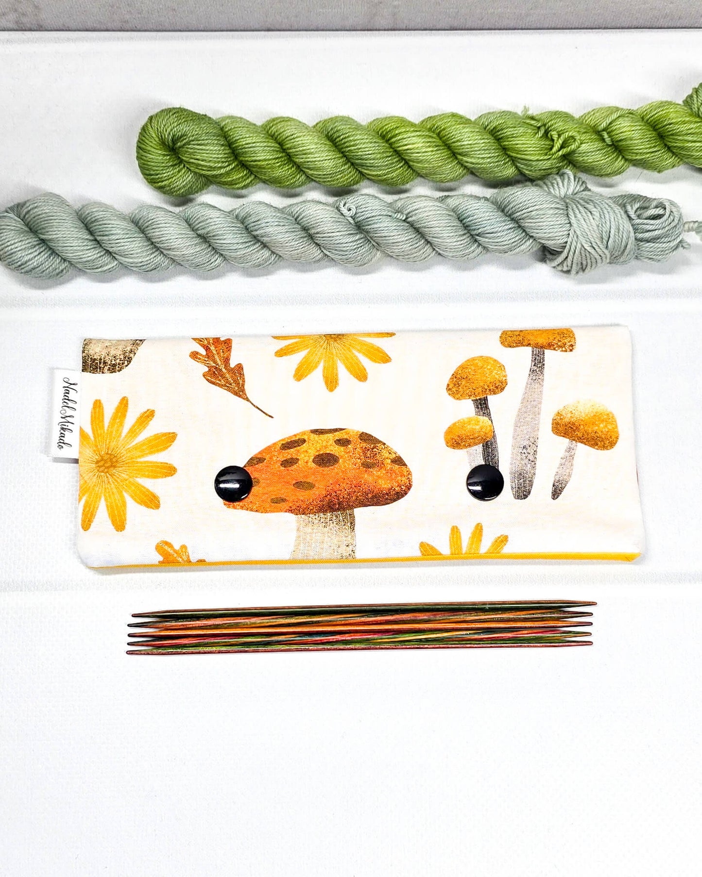 Nadelgarage für 15 cm lange Nadelspiele mit zwei schwarzen Druckknöpfen, verschiedenen Waldpilzen und gelben Blüten auf hellem Stoff. Handgenäht in Bayern von Nadelmikado.