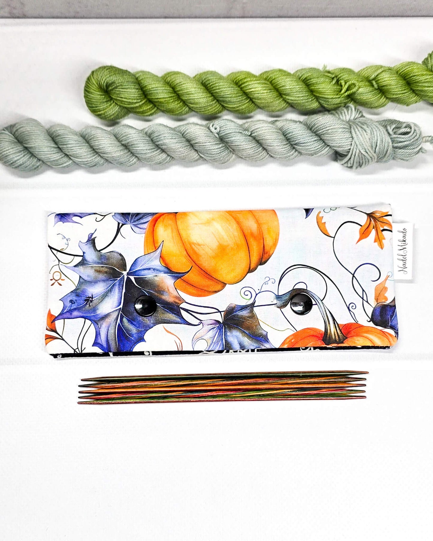 Nadelgarage für 15 cm lange Nadelspiele mit zwei schwarzen Druckknöpfen, großen orangefarbenen Kürbissen und blau-violetten Herbstblättern auf weißem Stoff. Handgenäht in Bayern von Nadelmikado.