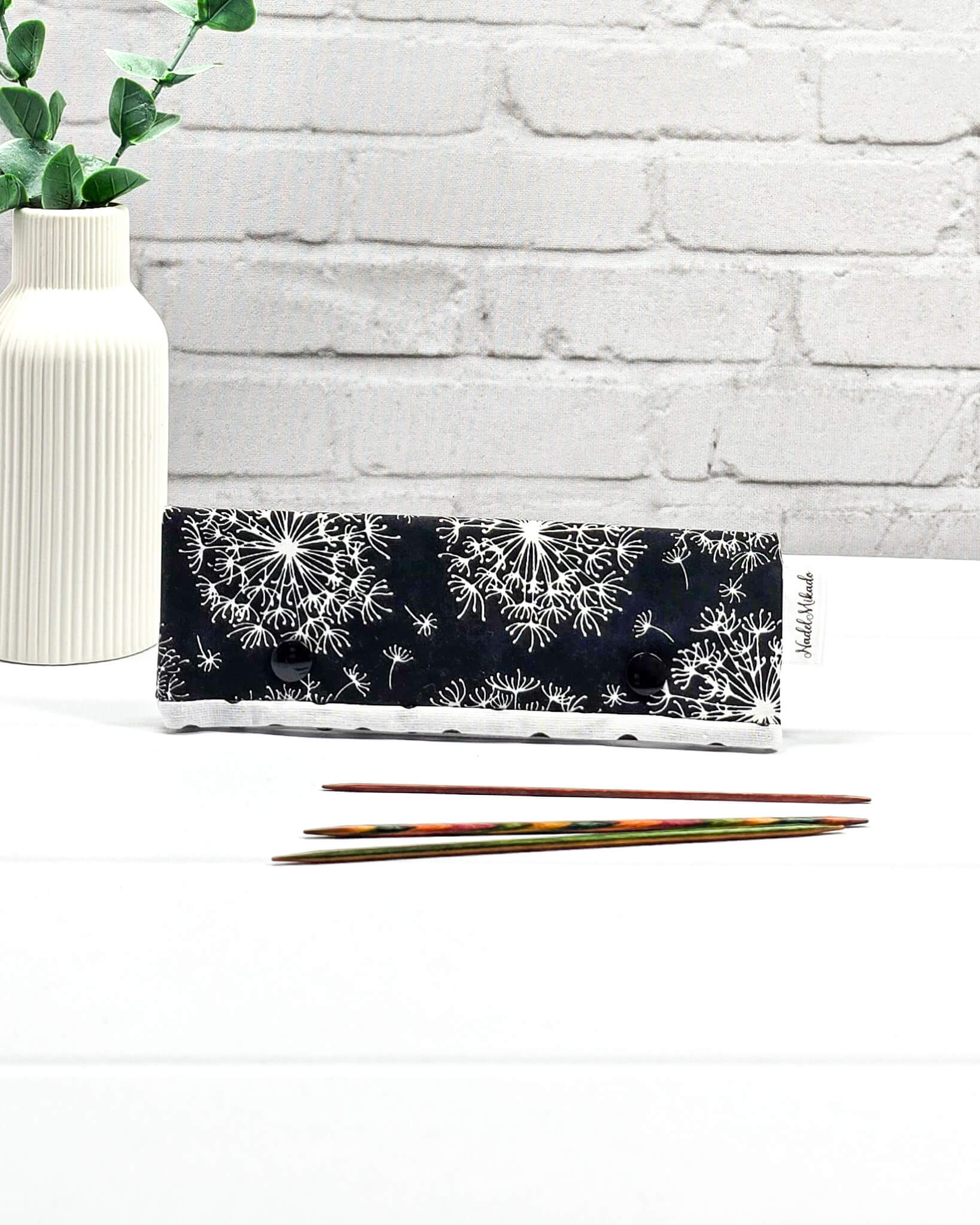 Nadelgarage 15cm aus schwarzem Stoff mit weißen Pusteblumen und zwei schwarzen Druckknöpfen, flach liegend vor drei kurzen Stricknadeln. Handgenäht in Bayern von Nadelmikado. 