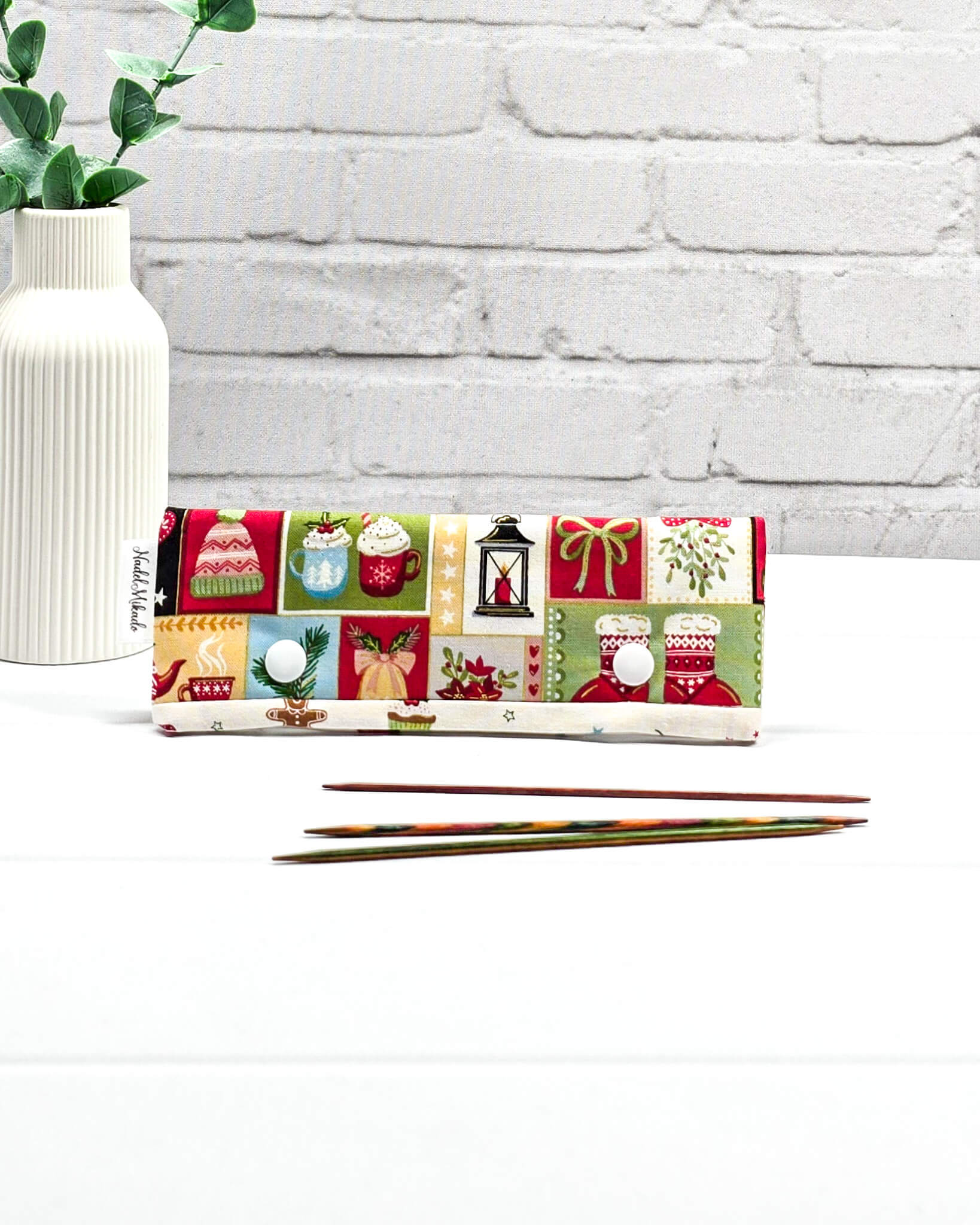 Nadelgarage 15cm mit bunten Weihnachtsmotiven und zwei weißen Druckknöpfen, flach liegend vor drei kurzen Stricknadeln. Handgenäht in Bayern von Nadelmikado