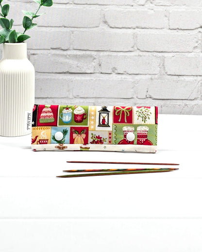 Nadelgarage 15cm mit bunten Weihnachtsmotiven und zwei weißen Druckknöpfen, flach liegend vor drei kurzen Stricknadeln. Handgenäht in Bayern von Nadelmikado