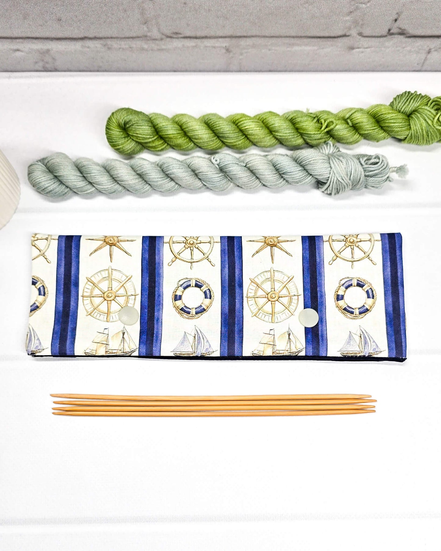 Nadelgarage für 20cm Nadelspiele aus cremeweißem Stoff mit blauen Längsstreifen, maritimen Motiven wie Steuerrädern und Schiffen sowie zwei hellen Druckknöpfen. Handgenäht in Bayern von Nadelmikado. 