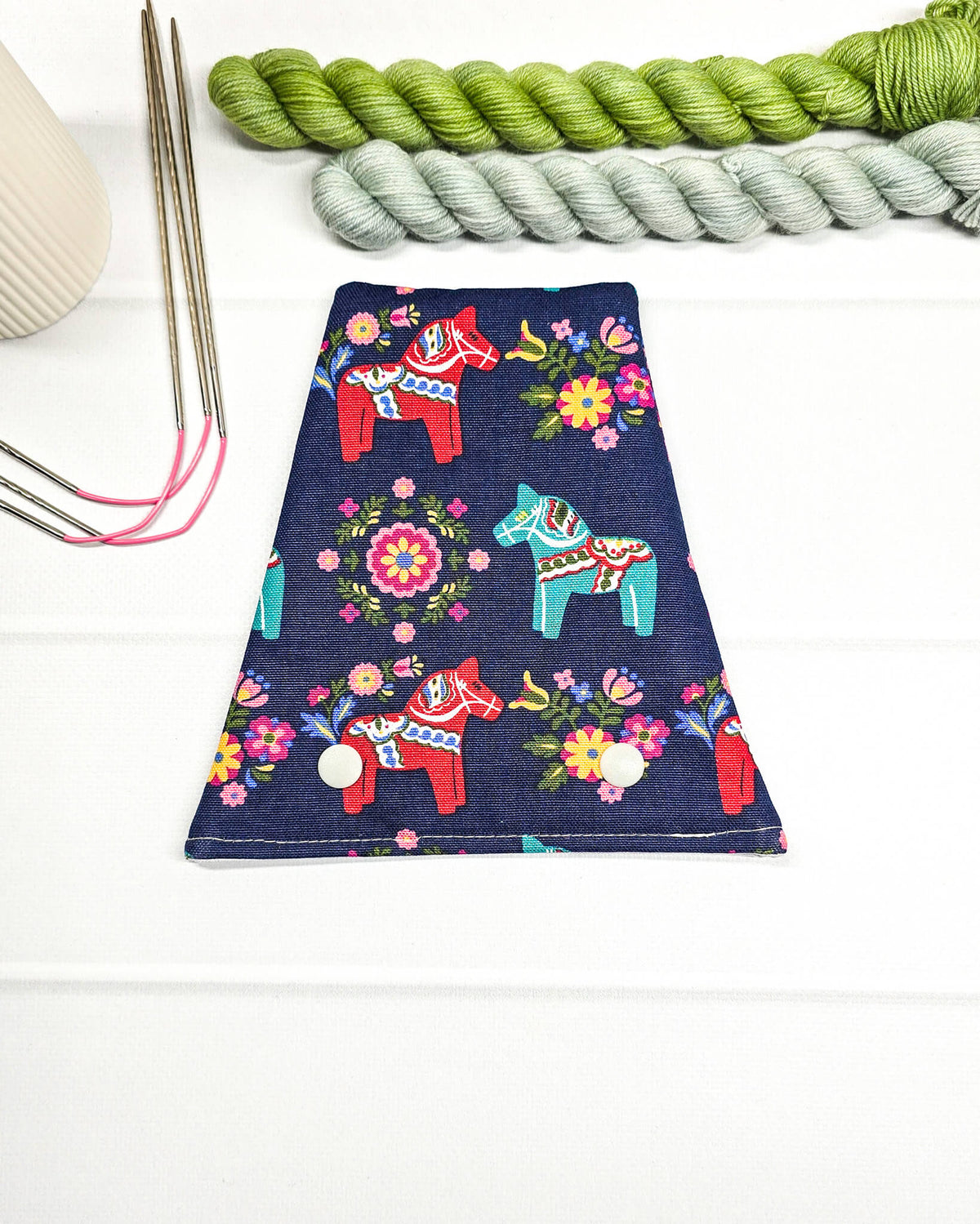 Nadelgarage für Trio-Stricknadeln aus dunkelblauem Stoff mit bunten Dala-Pferden, Blumenmotiven und zwei hellen Druckknöpfen. Handgenäht in Bayern von Nadelmikado.