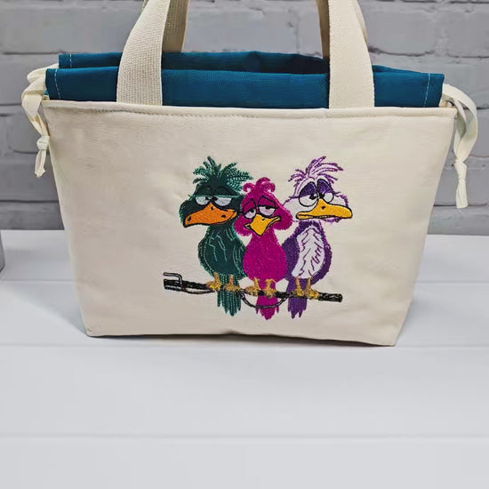 Projekttasche Alva S aus hellem Canvas mit Stickmotiv drei bunte Vögel auf Stange, Henkeln und petrolblauem Kordelzug. Handgenäht in Bayern von Nadelmikado.