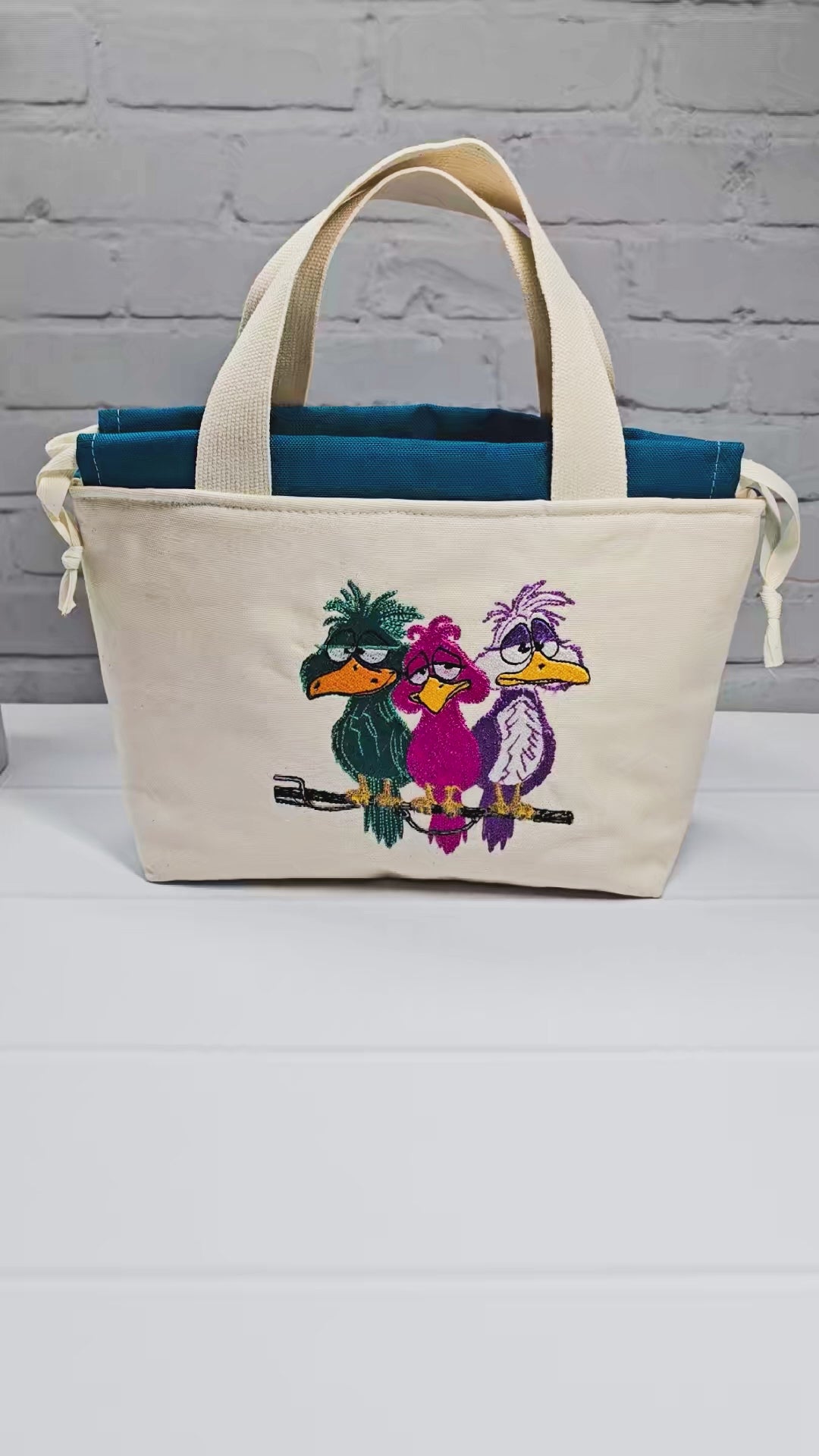 Projekttasche Alva S aus hellem Canvas mit Stickmotiv drei bunte Vögel auf Stange, Henkeln und petrolblauem Kordelzug. Handgenäht in Bayern von Nadelmikado.