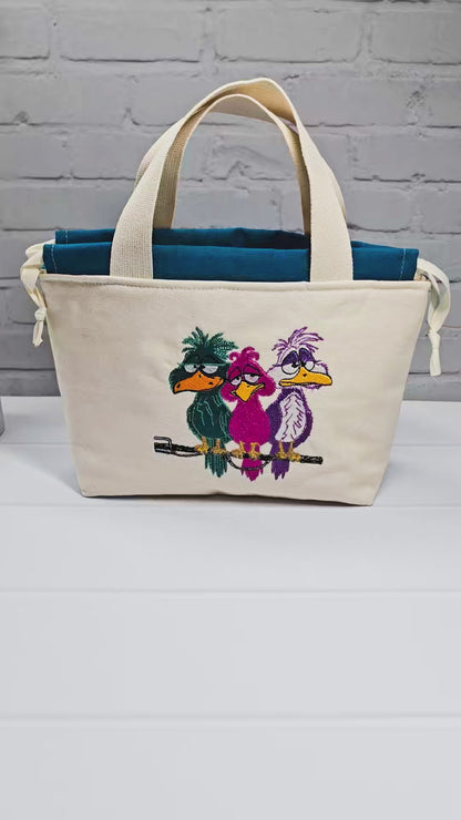 Projekttasche Alva S aus hellem Canvas mit Stickmotiv drei bunte Vögel auf Stange, Henkeln und petrolblauem Kordelzug. Handgenäht in Bayern von Nadelmikado.