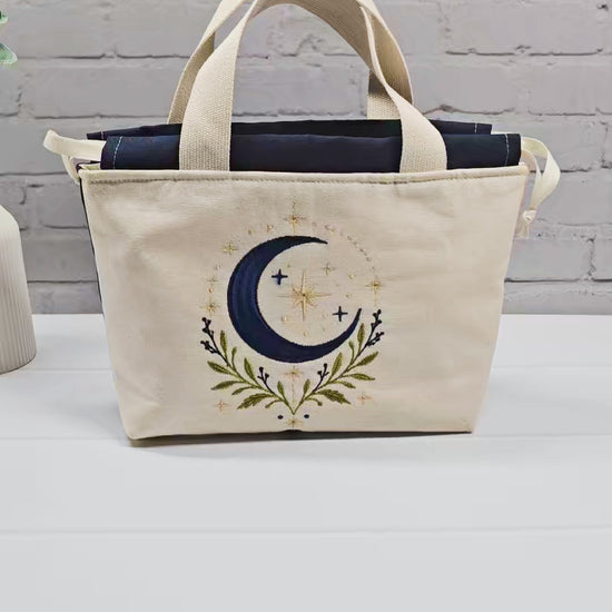 Projekttasche Alva S aus hellem Canvas mit Stickmotiv blaue Mondsichel und Sterne, Henkeln und dunkelblauem Kordelzug. Handgenäht in Bayern von Nadelmikado.