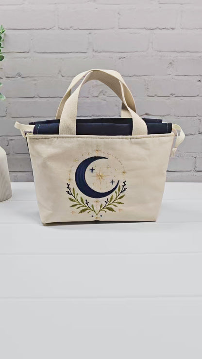 Projekttasche Alva S aus hellem Canvas mit Stickmotiv blaue Mondsichel und Sterne, Henkeln und dunkelblauem Kordelzug. Handgenäht in Bayern von Nadelmikado.