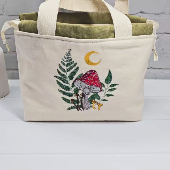 Projekttasche Alva S aus hellem Canvas mit Stickmotiv Fliegenpilz Farn und Mondsichel, Henkeln und olivgrünem Kordelzug. Handgenäht in Bayern von Nadelmikado.