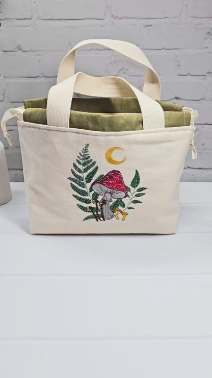 Projekttasche Alva S aus hellem Canvas mit Stickmotiv Fliegenpilz Farn und Mondsichel, Henkeln und olivgrünem Kordelzug. Handgenäht in Bayern von Nadelmikado.