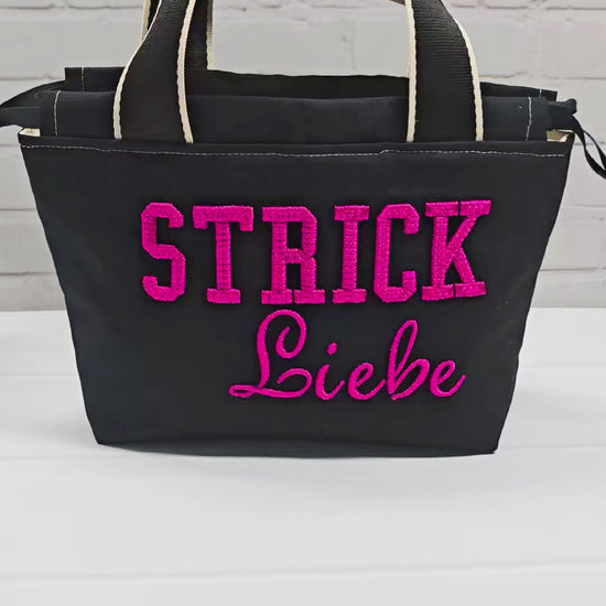 Projekttasche Alva S aus schwarzem Canvas mit erhabenem 3D-Stickzug Strickliebe in Pink, Henkeln und Kordelzugkragen. Handgenäht in Bayern von Nadelmikado.