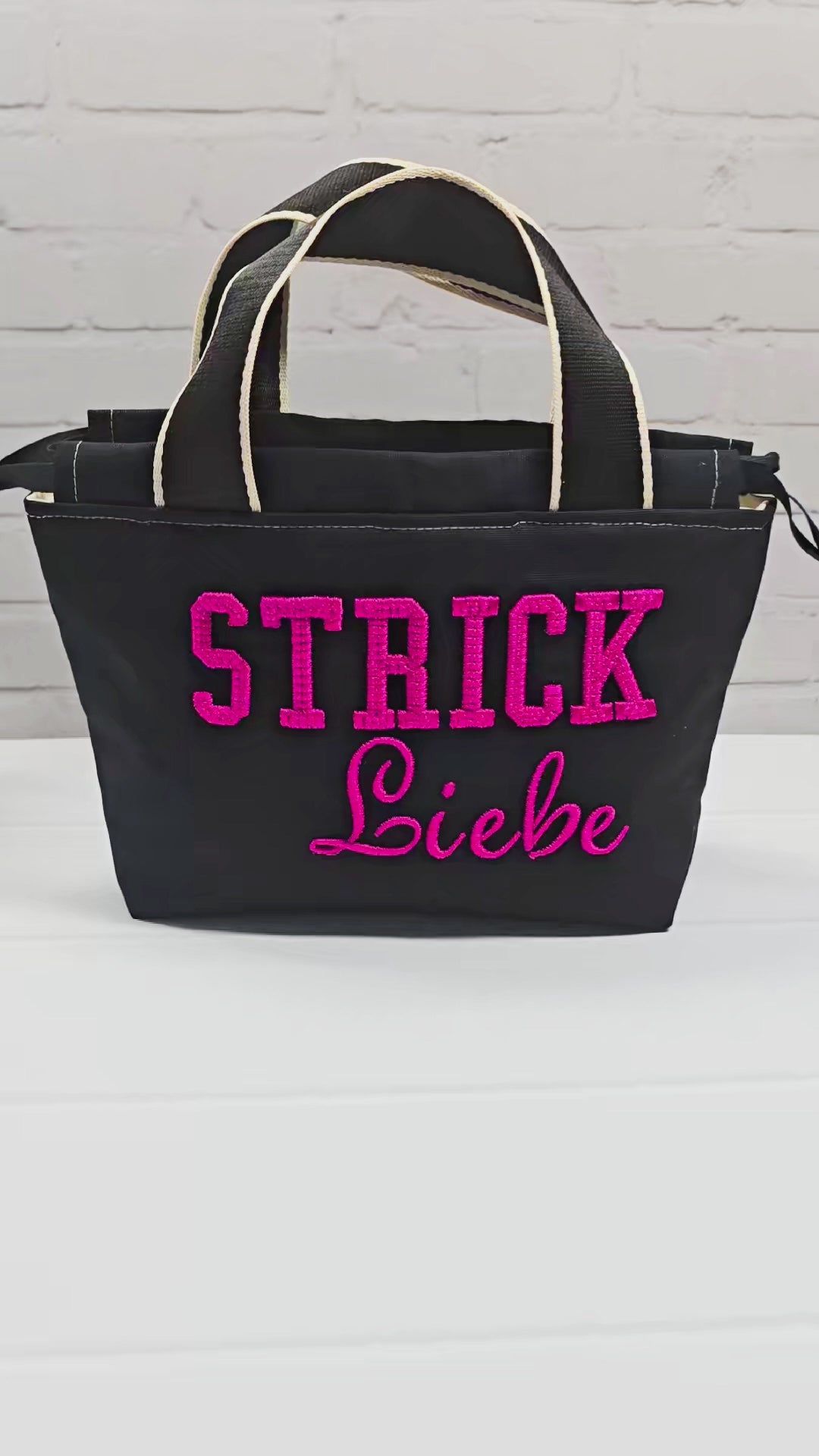 Projekttasche Alva S aus schwarzem Canvas mit erhabenem 3D-Stickzug Strickliebe in Pink, Henkeln und Kordelzugkragen. Handgenäht in Bayern von Nadelmikado.